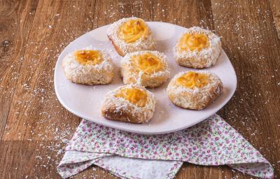 Foto da receita de pão com creme de leite moça, servidos em um prato rosa claro, com coco ralado salpicado, em uma bancada de madeira decorada com um pano de motivos florais