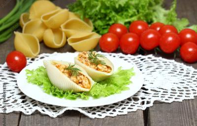 Fotografia de duas unidades de macarrão tipo concha recheado com creme de palmito sobre uma folha de alface, que está em um prato redondo branco e pequeno. Ao fundo, algumas unidades de macarrão tipo concha crus e ao lado, tomates cereja.