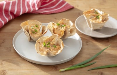 Fotografia em tons claros de um prato raso cinza claro com três tortilhas com frango. Ao lado tem outro prato raso, menor, com uma porção apenas. Os pratos estão sobre uma mesa de madeira clara. Fotografia em tons claros de um prato raso cinza claro com três tortilhas com frango. Ao lado tem outro prato raso, menor, com uma porção apenas. Os pratos estão sobre uma mesa de madeira clara.