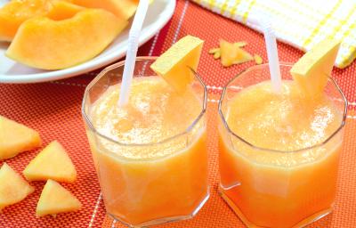 Fotografia de dois copos de vidro com vitamina de melão e maçã, com um pedaço de melão decorando do copo. Ao lado, pedaços de melão sobre uma toalha de mesa vermelha, e no fundo, um prato branco com fatias de melão.