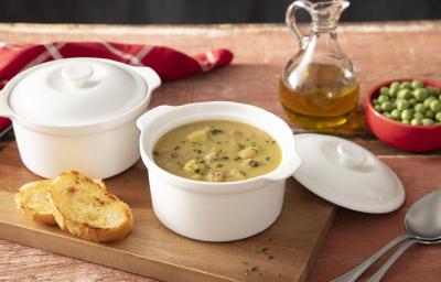 Fotografia em tons de marrom e vermelho de uma bancada de madeira, ao centro uma tábua com 2 mini caçarolas brancas, em uma delas, é possível ver o creme verde da sopa de ervilha. Ao fundo, azeite e ervilhas congeladas.