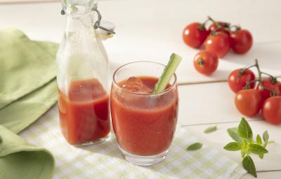 Fotografia em tons de vermelho em uma bancada de madeira clara, um pano verde liso e outro xadrez, um copo de vidro com o suco de tomate e uma jarra com mais suco ao centro da foto. Ao lado, tomates pequenos para decorar a mesa.