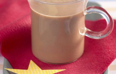 Fotografia em tons de vermelho e azul em uma bancada de madeira azul, uma toalhinha listrada em azul e rosa, um paninho vermelho com uma estrela amarela. Ao centro, uma xícara de vidro com chocolate quente.