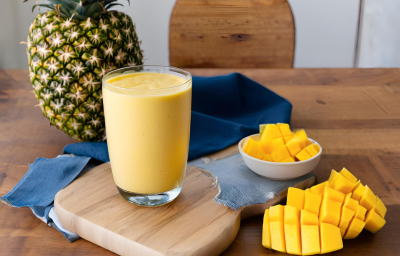 Fotografia de um copo de vidro com smoothie de iogurte, manga e abacaxi. O copo está sobre uma mini tábua de madeira, ao lado de dois recipientes brancos pequenos com pedaços de manga. No fundo tem um abacaxi inteiro sobre um pano azul escuro.