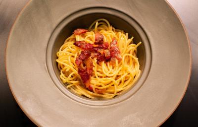 Fotografia em tons de amarelo com um prato cinza ao centro. Dentro do prato existe uma receita de macarrão carbonara finalizada com pedaços de guanciale frito.