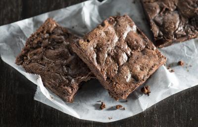 Fotografia de pedaços de brownie, em tom marrom, sob um papel branco e fundo preto.