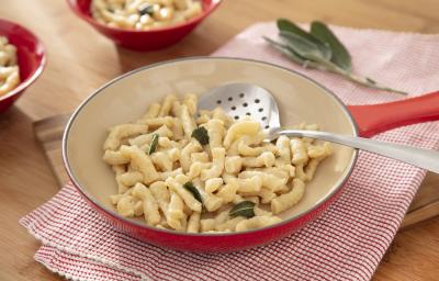 Foto da receita de Spatzle. Observa-se uma frigideira grande sobre um pano de prato quadriculado vermelho. A massa está dentro da frigideira com folhas de manjericão.