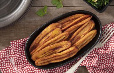 Fotografia em tons de vermelho em uma bancada de madeira, uma toalha vermelha com flores, um recipiente oval preto com várias bananas fritas dentro dele.