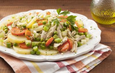 Fotografia em tons de marrom e branco de uma bancada de madeira com um paninho branco com listras vermelhas e amarelas sobre ele um prato branco redondo com bordas irregulares e a salada de frango com legumes. Ao lado um recipiente de vidro com azeite.