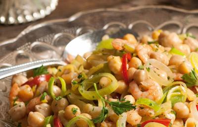 Fotografia em tons de bege e marrom, com salada de grã0-de-bico e legumes de cores verde e vermelha, dentro de saladeira de vidro com uma colher de servir, tudo sobre bancada de madeira.