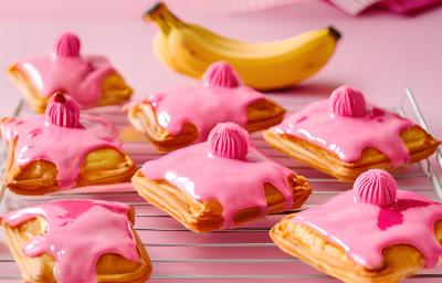 Fotografia de vários pasteis pequenos quadrados, recheados de banana e leite ninho, com cobertura de creme de nesquik. Os pasteis estão sobre uma grande branca, sobre uma mesa rosa.