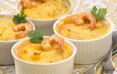 Fotografia em tons de branco e amarelo de uma bancada de madeira marrom, um paninho cinza e um prato amarelo com branco, sobre ele quatro recipientes redondo com o escondidinho de camarão decorados com um camarão e uma folha de salsinha.