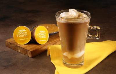 Fotografia em tons de amarelo em uma bancada de madeira escura, um paninho amarelo, uma xícara de vidro alta com o Latte Macchiato Dolce Gusto dentro, com doce de leite e decorado com chantilly. Ao fundo, cápsulas do café e um potinho com doce de leite.