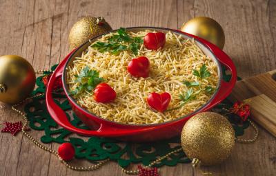 Fotografia em tons de vermelho em uma bancada de madeira, um pano redondo natalino verde, um prato vermelho redondo grande e fundo com a salada de salpicão coberta com batata palha e tomate-cereja dentro dele. Ao lado, enfeites de Natal.