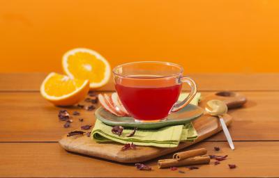 Fotografia em tons de laranja em uma bancada de madeira e um fundo laranja. Ao centro, uma tábua de madeira, um paninho verde e uma xícara de vidro com o chá de hibisco dentro dele. Ao fundo, laranja e canela em pau espalhados pela bancada.