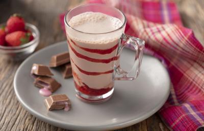 fotografia em tons de marrom, branco, vermelho e rosa de uma bancada marrom vista de frente. Contém um prato redondo branco, com um copo transparente que contém a bebida de morango, e ao lado cubos de chocolate com recheio de morango.