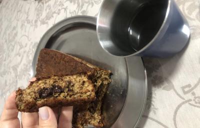 Fotografia de uma mão feminina segurando um pedaço de bolo de banana com aveia e chocolate. A outra parte do bolo está atrás da mão, sobre um recipiente oval de alumínio, ao lado de um copo de aluminio com café.