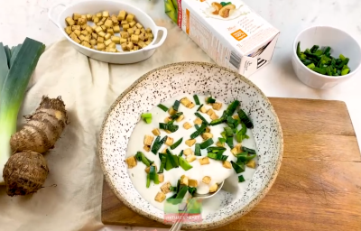 Foto vista de cima de uma mesa com a receita de creme de alho poró com inhame, num prato de cor clara, sobre uma tábua de madeira. Na mesa há potes com croutons, ervas e uma embalagem de bebida vegetal Nature's Heart