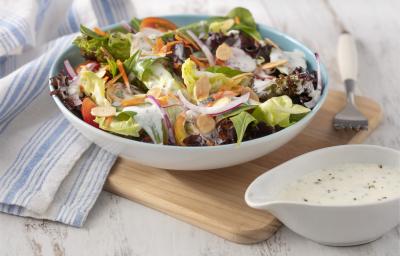 Fotografia em tons de branco e azul de uma bancada branca e uma tábua de madeira, sobre ela um recipiente redondo com a salada e um garfo. Ao lado uma molheira branca com o molho e um paninho branco e azul.