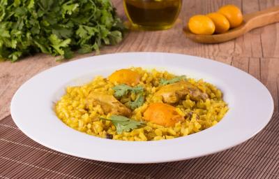 Fotografia em tons de amarelo, em uma bancada de madeira de cor marrom. Ao centro, um prato branco redondo contendo o arroz com frango e pequi. Ao fundo, uma colher de pau contendo alguns pequis e ao lado alguns temperos verdes espalhados.