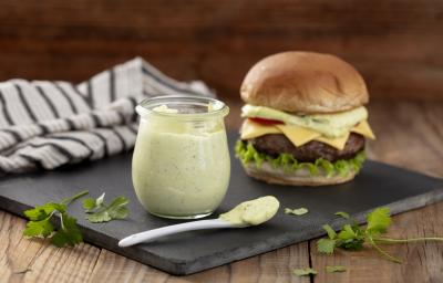 Fotografia em tons de verde em uma bancada de madeira clara, um pano branco com listras pretas, uma tábua de ferro preta e em cima dela, um potinho de vidro com a maionese verde caseira e ao lado um hambúrguer para acompanhar essa deliciosa maionese.