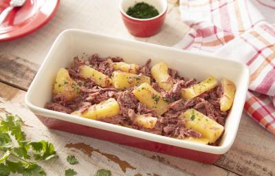 Fotografia em tons de vermelho de uma bancada de madeira, ao centro uma travessa vermelha por fora e branca por dentro com pedaços de mandioca e carne-seca desfiada. Ao redor, folhas de salsa e um paninho quadriculado branco e vermelho.