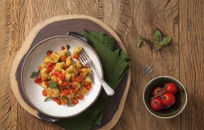 fotografia em tons de marrom e verde de uma bancada marrom vista de cima. Contém um recipiente branco e redondo com Nhoque de Abóbora e molho de tomate com um garfo ao lado para servir. Abaixo um pano verde e ao lado um pote redondo com tomates.