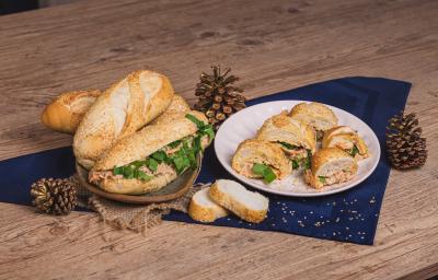 Fotografia em tons de azul escuro em uma bancada de madeira, um pano azul escuro, pinhas natalinas e um prato branco ao centro com fatias do pão com gergelim recheado de atum, cenoura, creme de ricota e rúcula. Ao lado, uma cesta de pães.