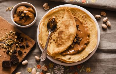 Fotografia de crepes recheados com doce de leite e calda de gengibre sobre um prato de vidro branco e uma colher de sobremesa em cima. Ao redor, uma tábua de madeira, pistaches e chocolate espalhados e doce de leite dentro de um pote pequeno de vidro.