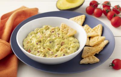 Fotografia em tons de azul em uma bancada de madeira clara com um pano laranja ao lado e um prato grande azul e um prato fundo branco com o guacamole dentro. Ao lado, um pedaço de avocado, alguns tomatinhos e biscoitinhos.