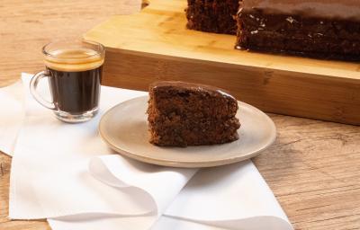 Fotografia de uma bancada de madeira, e sobre ela tem uma torta de chocolate com café em um prato de vidro em tom pastel, que está apoiado sobre um pano branco. Ao lado da sobremesa tem uma xícara com Nescafé, também sobre o pano.