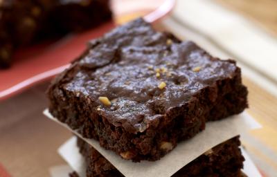 Fotografia em tons de marrom e branco de uma bancada de madeira clara e um paninho marrom, sobre ela fatias de brownie separadas por papel manteiga. Ao fundo um prato vermelho com pedaços de brownie.