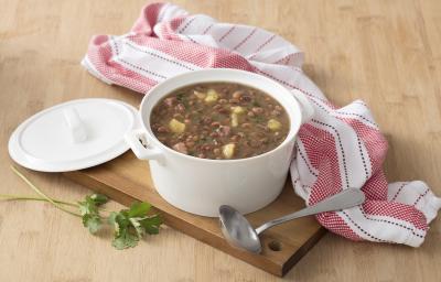 Fotografia em tons de marrom, branco e rosa de uma bancada de madeira marrom vista de cima, contém uma tábua de madeira com uma panela redonda branca com alças e feijão dentro com pedaços de carne-seca e mandioca. Ao lado a tampa da panela e um pano.