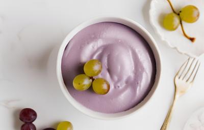 Fotografia em tons de branco e roxo com um pote branco ao centro. Dentro do pote existe uma receita de mousse de uva de cor roxa com algumas uvas verdes de enfeite.