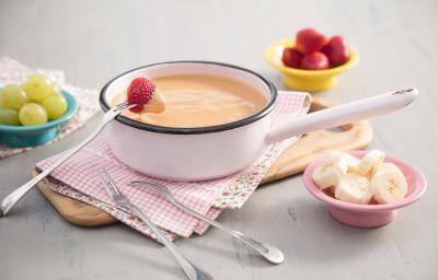 Fotografia em tons de branco, caramelo e rosa, tendo ao centro panela em ágata contendo doce de leite cremoso, garfinhos para fondue ao lado sobre guardanapo quadriculado e peça de madeira, no entorno potinhos com frutas picadas, tudo sobre bancada cinza.