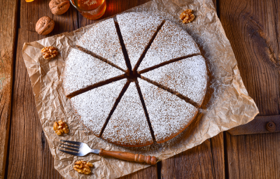 Foto em tons de marrom da receita de bolo alemão de mel servida cortada em 8 pedaços polvilhada com açúcar de confeiteiro, bolo em cima de uma mesa de madeira com nozes ao lado