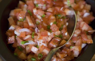 Fotografia de um molho vinagrete com cebola e salsinha, e uma colher apoiada na salada. A vinagrete está em um recipiente pequeno e preto.