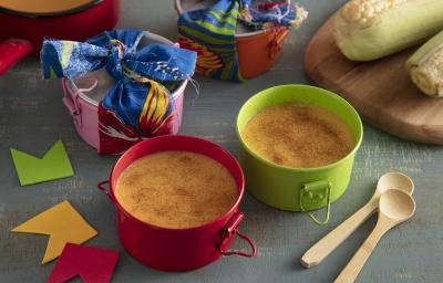 Foto da receita de Curau Leve. Observa-se duas panelinhas como curau polvilhado de canela. A foto é decorada com bandeirinhas de festa junina, com milhos verdes e colherinhas de madeira