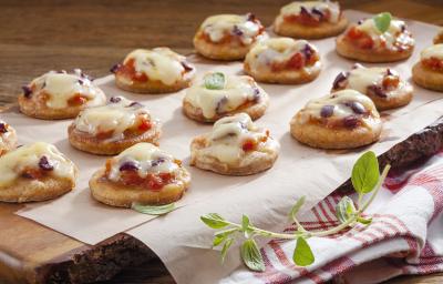 Fotografia em tons de marrom em uma bancada de madeira com uma tábua de madeira escura ao centro com muitas mini pizzas de frigideira apoiadas nela. Embaixo, um pano branco com listras vermelhas.