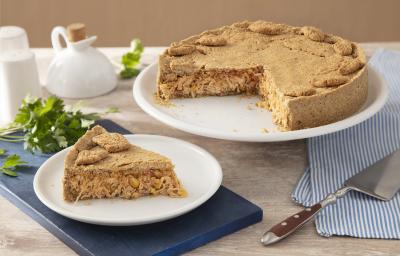 Receita da Torta de Frango sem Glúten. Observa-se a torta em um prato grande branco e, ao lado esquerdo, um prato individual com uma fatia. Como decoração da foto, tem-se um guardanapo listrado azul e uma espátula de cortar.