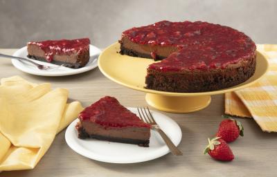 Fotografia em tons de amarelo em uma bancada de madeira com um suporte para bolo amarelo ao centro e a torta cheesecake de chocolate em cima. Ao lado, um paninho xadrez amarelo e dois pratos brancos com duas fatias da cheesecake de chocolate em cada um.