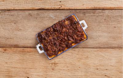 Fotografia em tons de marrom em uma bancada de madeira rústica com um recipiente azul com alças brancas laterais e a polenta cremosa coberta com molho de carne dentro do recipiente.