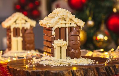 Imagem da receita de Casinha de KitKat Papai Noel, montada num formato miniatura de uma casinha de chocolate em tons marrom e branco, com um Papai Noel de chocolate à frente, sobre uma tábua de madeira e com decorações de natal ao redor