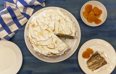 Fotografia em tons de azul e laranja em uma bancada azul, com um pano xadrez azul, um prato branco redondo com a torta de nozes em cima dele. Ao lado, um prato menor com a fatia da torta e damascos ao lado dele.