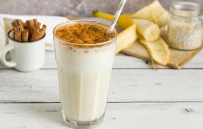 Fotografia de um copo de vidro com uma vitamina de banana com canela polvilhada e um canudo branco e cinza dentro do copo. Está sobre uma mesa de madeira branca, ao fundo tem paus de canela em um copinho branco e metade de uma banana aberta. Fotografia de um copo de vidro com uma vitamina de banana com canela polvilhada e um canudo branco e cinza dentro do copo. Está sobre uma mesa de madeira branca, ao fundo tem paus de canela em um copinho branco e metade de uma banana aberta.