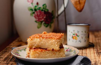 Foto da receita de Bolo de Arroz Cozido. Observa-se 3 fatias retangulares de bolo empilhadas e, atrás, um cafézinho para acompanhar.