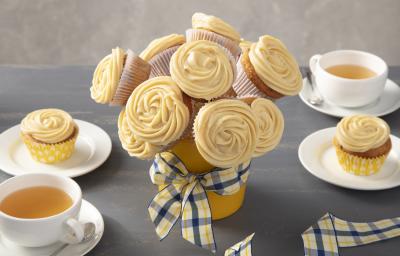 Fotografia em tons de amarelo em uma bancada cinza com um vaso de buquê de cupcake ao centro com um laço xadrez em amarelo e azul. Ao lado, xícaras brancas com chá e pratos brancos com um cupcake em cada um.