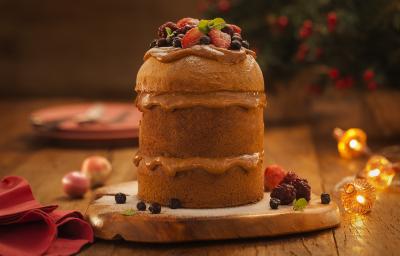 Foto de um panettone cortado em três camadas, com um recheio em tom de marrom claro. Ele está sobre uma tábua de madeira há decorações de natal