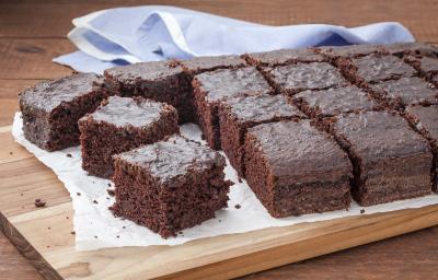 16 pedaços de bolo de chocolate com calda em cima de uma toalha branca que está em cima de uma tábua de madeira. Todos do mesmo tamanho