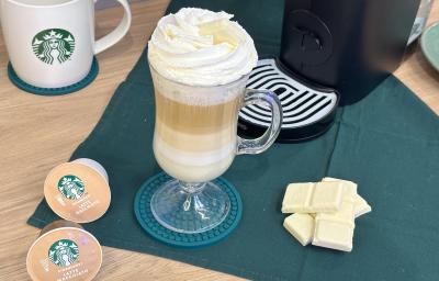 Foto da receita de Mocha Branco. Observa-se uma xícara transparente com a bebida em fases decorada com chantilly e, ao lado, cápsulas de Starbucks e quadradinhos de Galak.